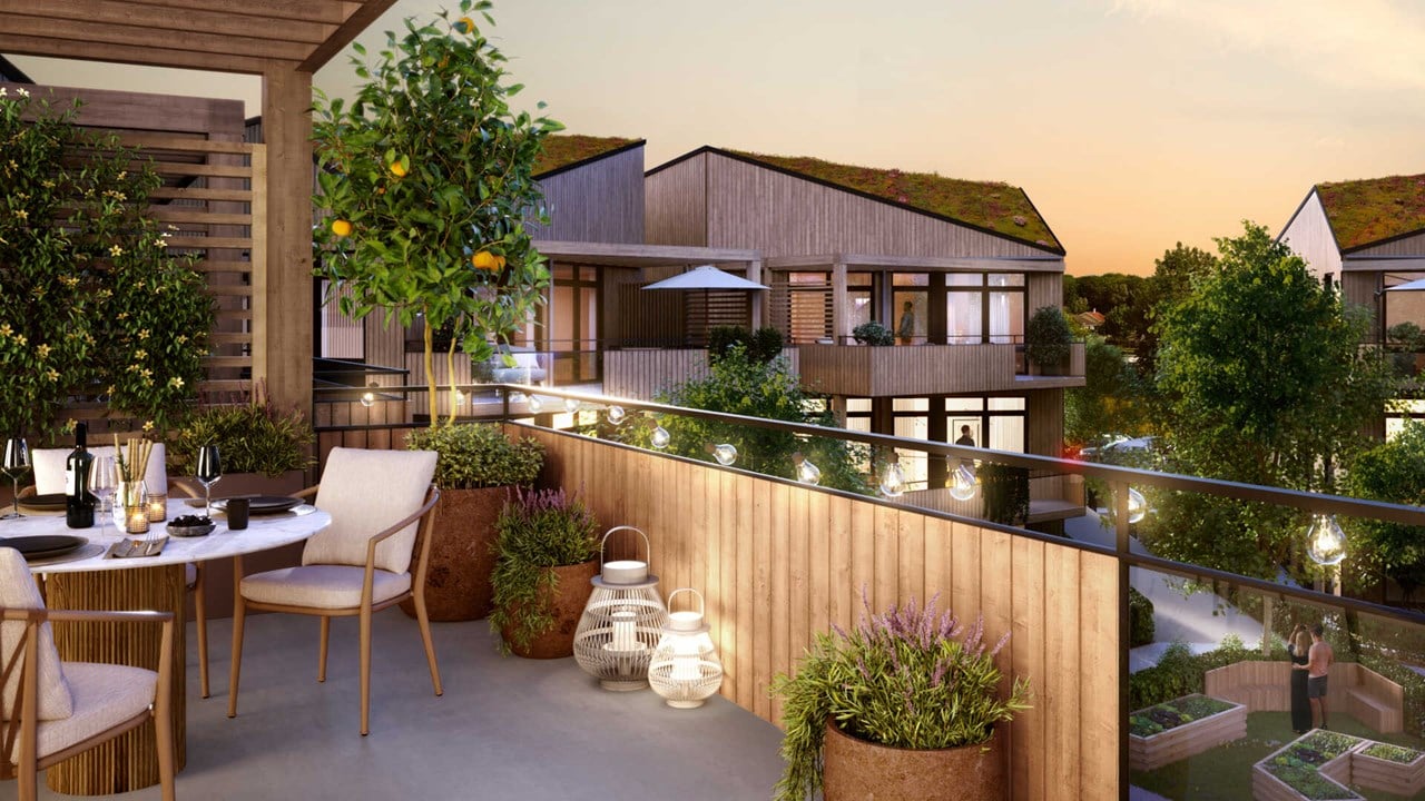 Balkong med bord och stolar, omgiven av växter, med utsikt över moderna trähus i kvällssken