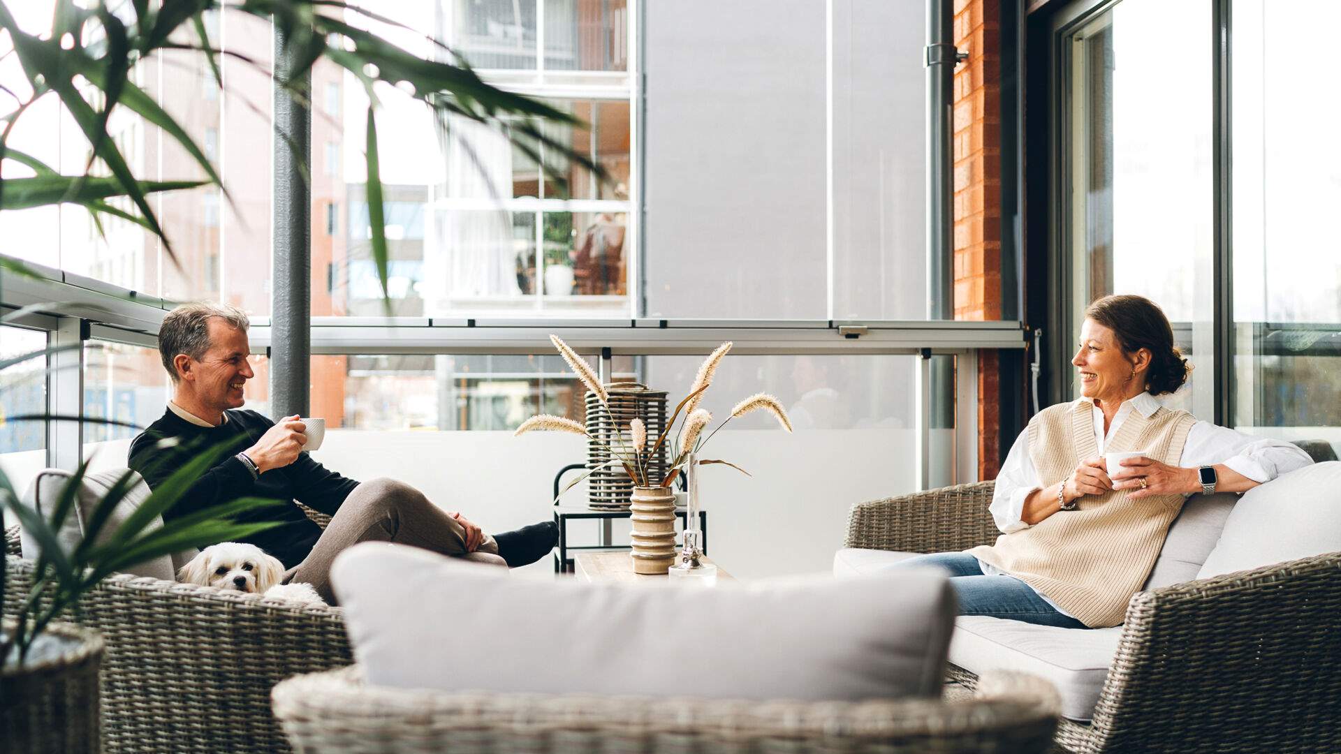 Två personer sitter i korgstolar på en inglasad balkong och dricker te eller kaffe, med en hund bredvid dem och växter i förgrunden