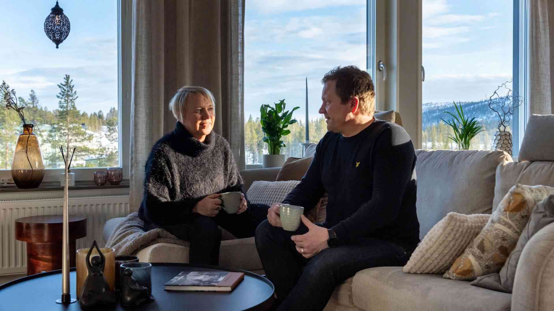 Man och kvinna sitter i en soffa med kaffekoppar, ett vinterlandskap syns genom fönstren