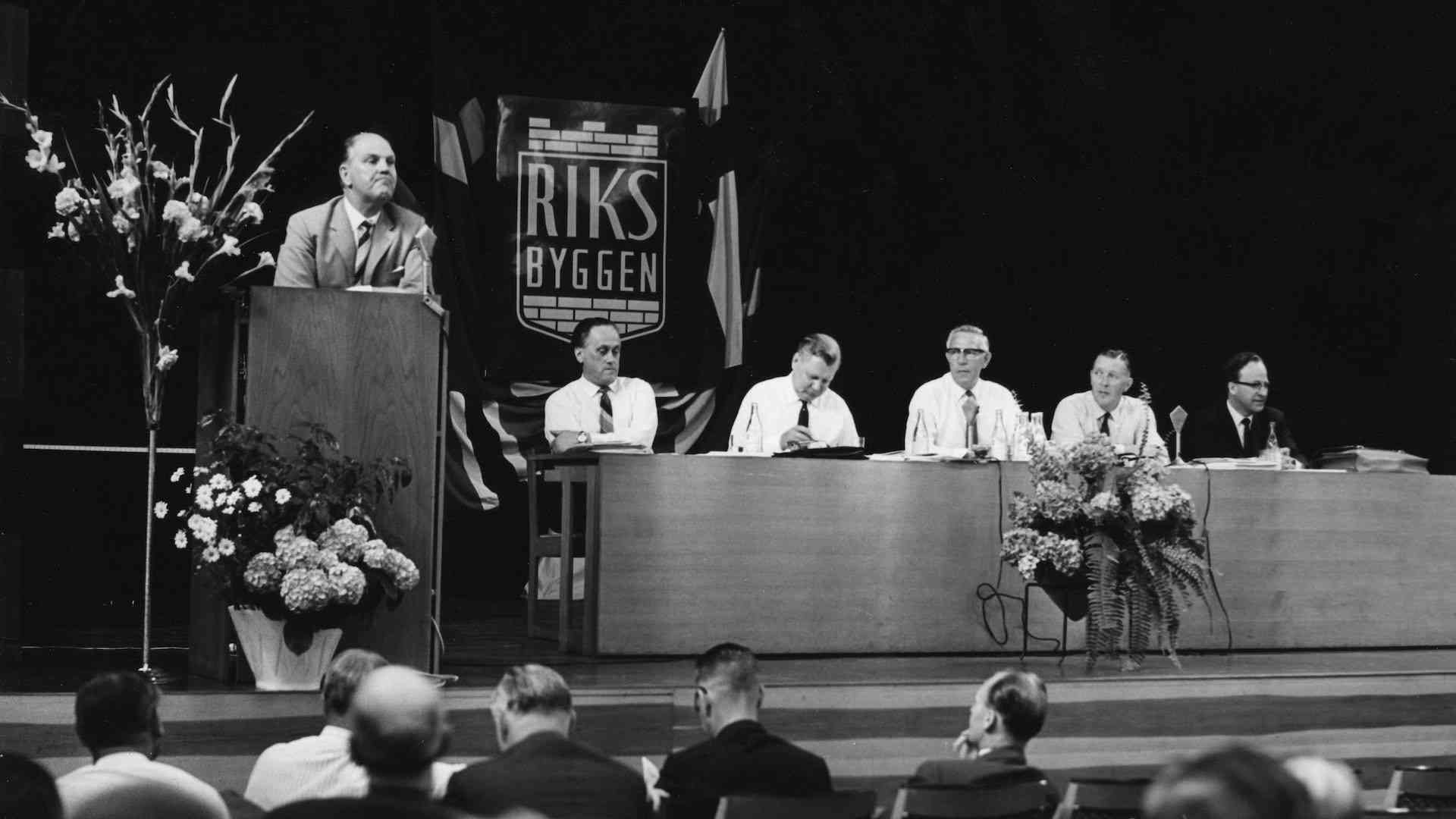 En man står vid ett podium och håller ett tal medan fem personer sitter vid ett bord på en konferens med Riksbyggen-logotypen i bakgrunden