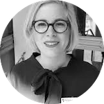 Kvinna med blont hår och glasögon, klädd i en mörk blus med rosett