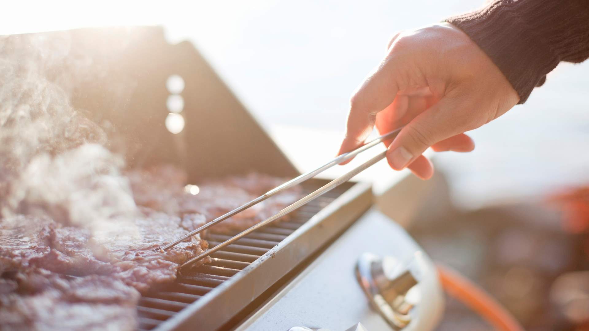 Hand som håller i grilltång och grillar kött på en utomhusgrill medan solen lyser in från vänster