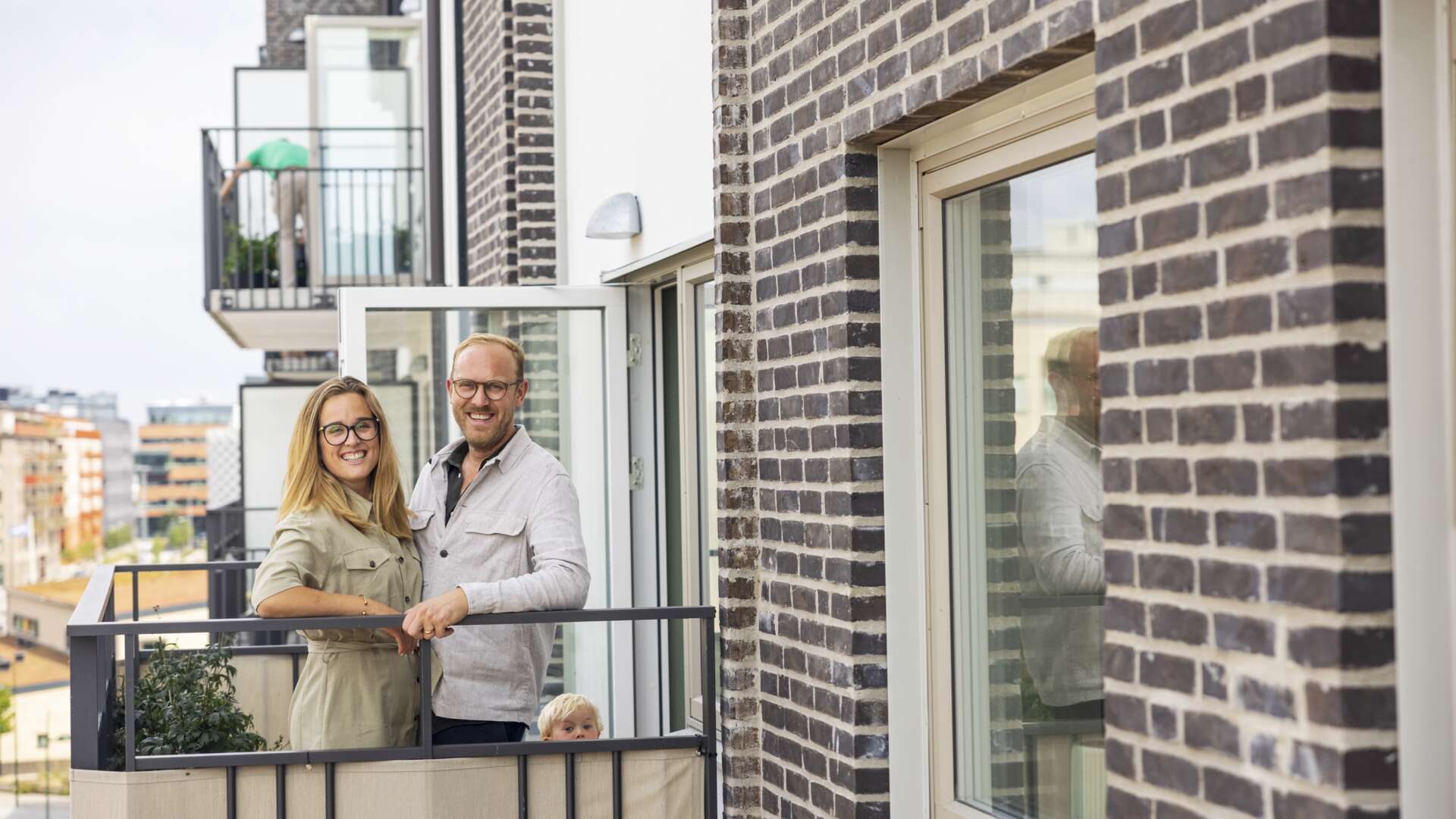 Personer står på en balkong med tegelväggar i bakgrunden. Ett barn tittar fram bakom kanten av balkongen