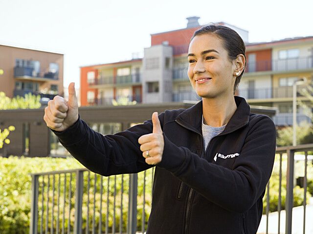Person som står utomhus framför flerbostadshus och visar tummen upp med båda händerna, klädd i en mörk tröja