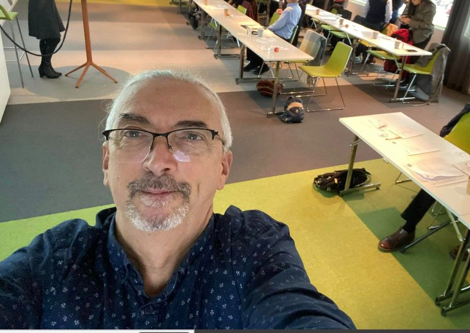 En konferens- eller möteslokal med deltagare som sitter vid bord, en person står vid ett podie och det finns spridda dokument och flaskor på borden