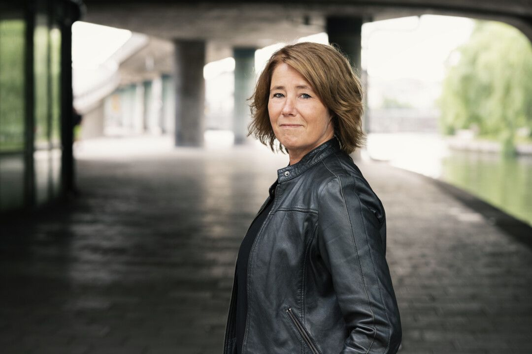 Person stående under en bro i en stadsmiljö, iklädd en svart läderjacka med en flod och grönska i bakgrunden