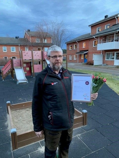 En person står på en lekplats med en certifikatsram i ena handen och en bukett rosa blommor i den andra. I bakgrunden syns flerbostadshus och en sandlåda