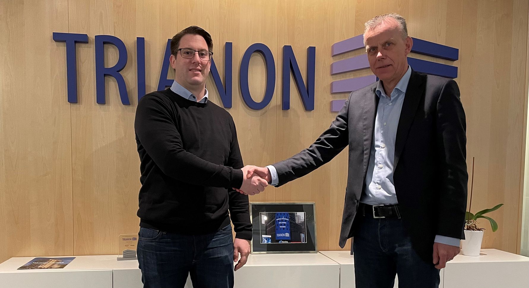 Två män skakar hand framför en vägg med texten "TRIANON" och en hylla med en liten skärm och en krukväxt