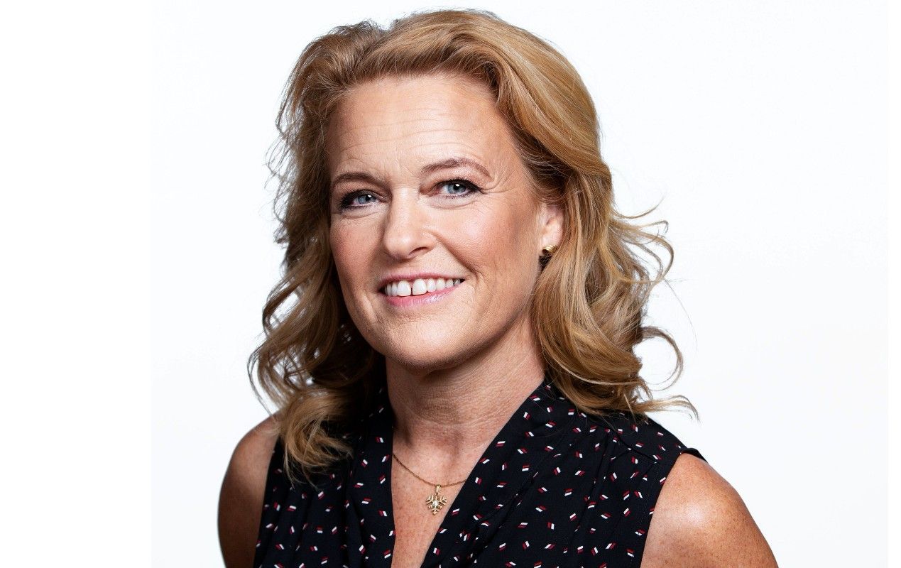 Kvinna med blont lockigt hår, iklädd en svart ärmlös blus med ett litet mönster och ett halsband med guldberlock