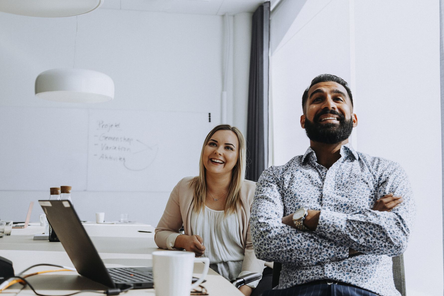 Två personer sitter vid ett skrivbord i ett modernt kontor med en bärbar dator, kaffekoppar och en whiteboard i bakgrunden