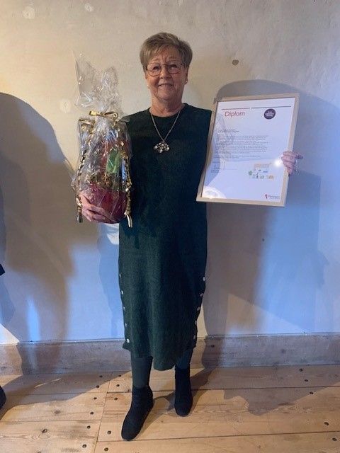 Person i mörkgrön klänning som håller i en inslagen present och ett diplom, stående på ett trägolv mot en ljus vägg