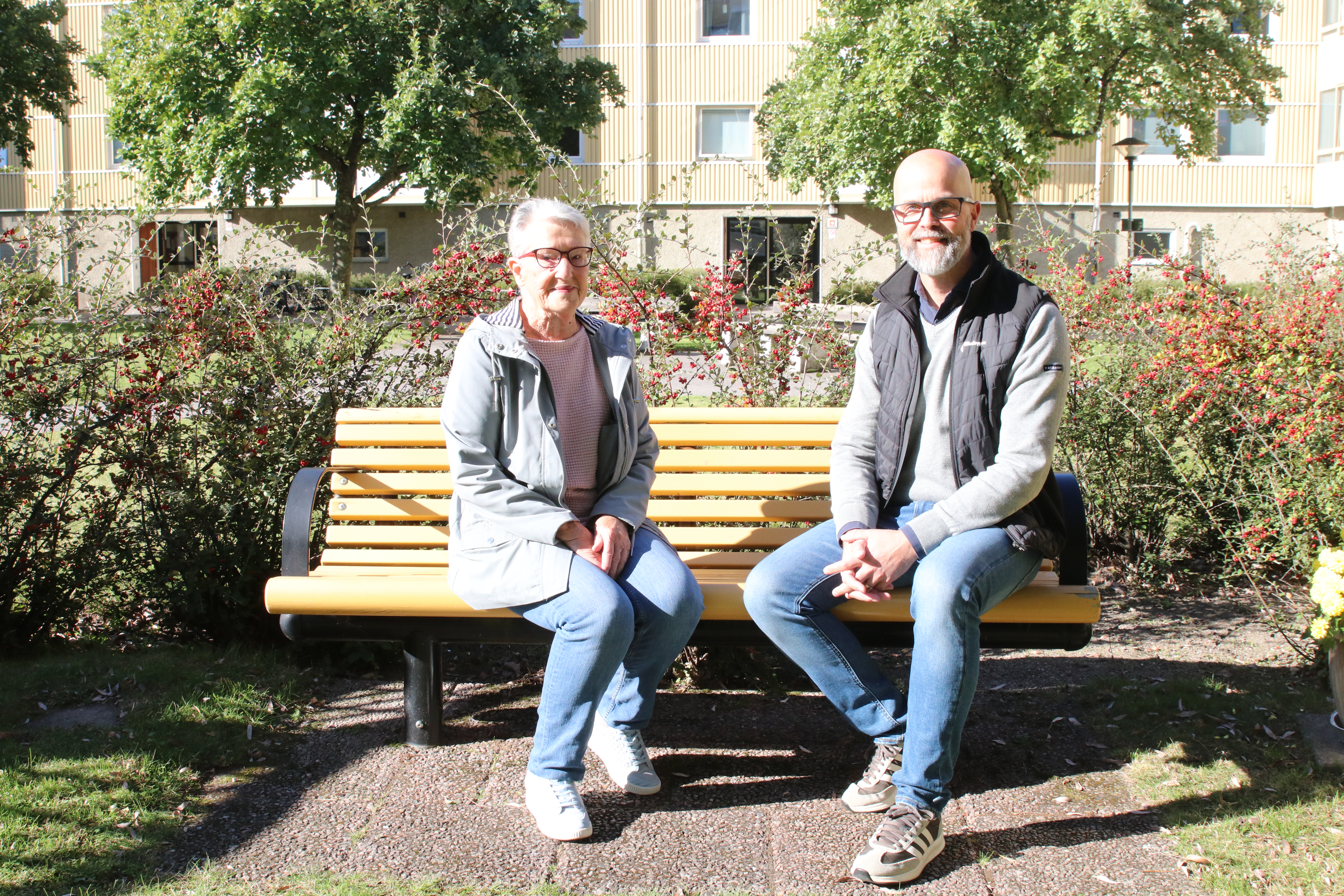 Äldre kvinna och man sitter på en bänk i en trädgård med grönt gräs och buskar i bakgrunden