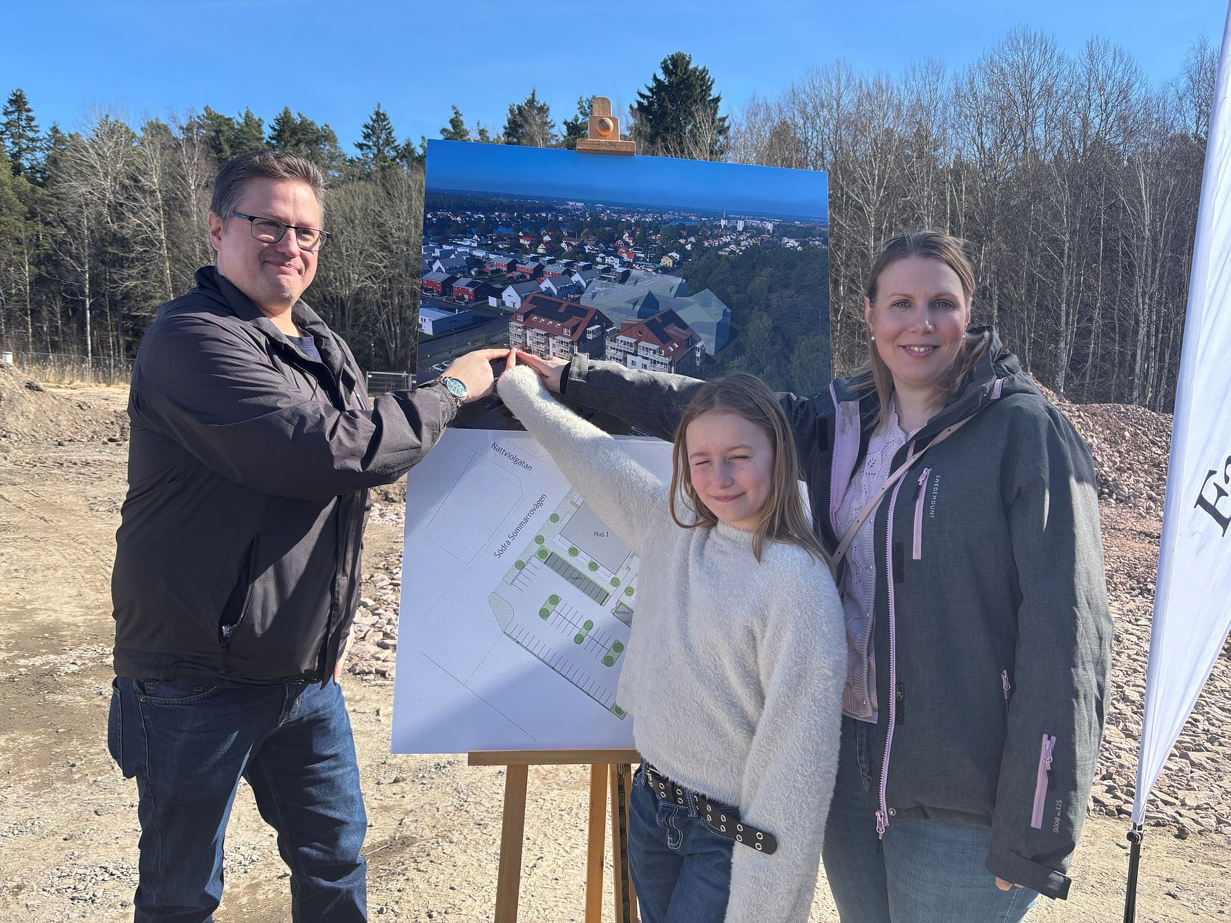 Tre personer pekar på en byggskylt utomhus vid en tomt, med ett flygfoto och en situationsplan