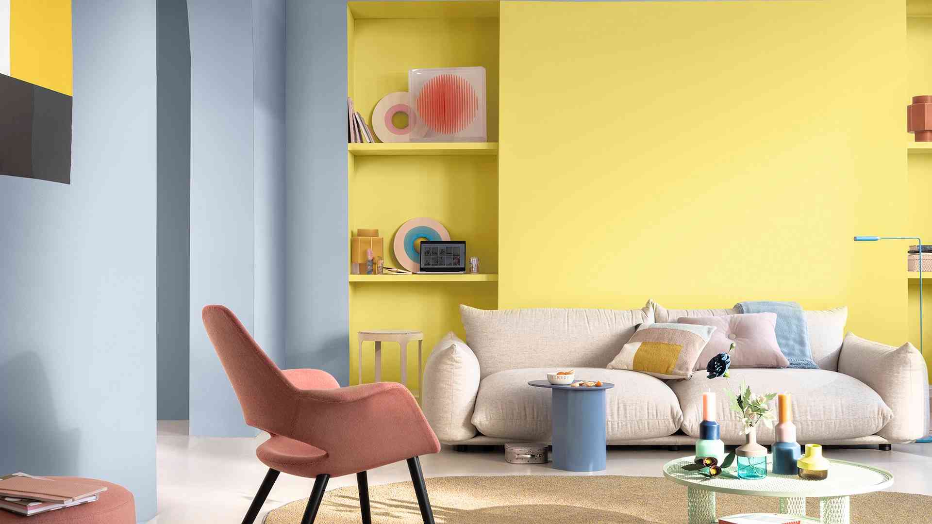 Färgglad vardagsrum med ljusblå och gula väggar, en beige soffa, rosa stol, och moderna hyllor med prydnadssaker