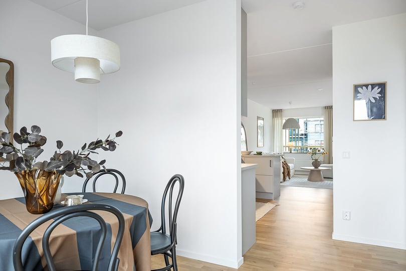Matplats med svartmålade stolar, rund bord med beige och brun duk, brun vas med dekorativa grenar, och vit taklampa i ett ljust rum. Bakgrunden visar ett öppet vardagsrum med stora fönster