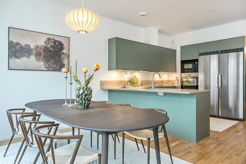 Modernt kök och matplats med gröna skåp, köksö, svart matbord, trästolar och tulpaner i en vas. Väggkonst och hängande lampa syns ovanför bordet