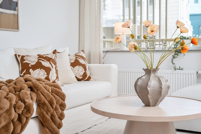 Soffgrupp med vita möbler, bruna textilier och orange blommor i en vas på ett runt soffbord. Ljusa gardiner och ett fönster med stadsvy i bakgrunden