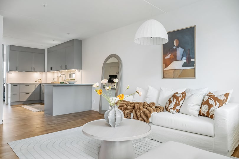 Ljust vardagsrum med vit soffa, blommig kudde, rund soffbord och vas med blommor, i ett öppet kök med grå skåp i bakgrunden